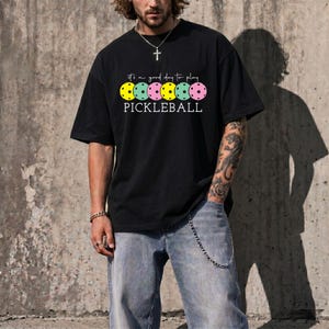 A man wearing a black t-shirt with a graphic design of colorful bowling pins and the text "It's a good day to play PICKLEBALL" printed on it.