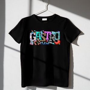 K&ouml;nnte beinhalten: Schwarzes T-Shirt mit einem bunten Grafikdesign, auf dem "Gastro Crew" steht. Die Grafik besteht aus verschiedenen medizinischen Bildern, darunter ein Stethoskop, Pillen und ein Magen.