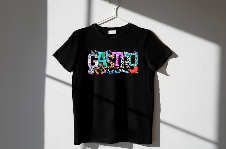 K&ouml;nnte beinhalten: Schwarzes T-Shirt mit einem bunten Grafikdesign, auf dem "Gastro Crew" steht. Die Grafik besteht aus verschiedenen medizinischen Bildern, darunter ein Stethoskop, Pillen und ein Magen.
