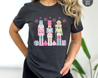 Peut inclure: Un t-shirt à manches courtes gris foncé chiné avec une impression graphique de trois casse-noisettes dans des tons rose et bleu pastel, entourés de flocons de neige, de cadeaux et de boules roses. Le texte "heather dark grey" est visible.