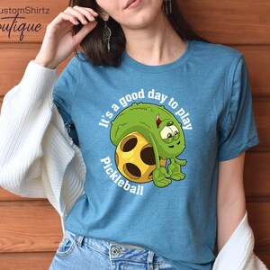 May include: A teal blue t-shirt with a cartoon pickleball character and the text "It's a good day to play pickleball".
