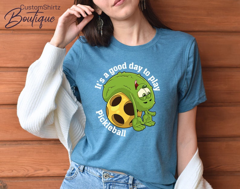 May include: A teal blue t-shirt with a cartoon pickleball character and the text "It's a good day to play pickleball".