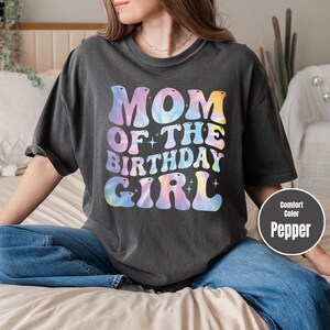 A woman wearing a black t-shirt with the text "MOM OF THE BIRTHDAY GIRL" printed on it, sitting on a bed.