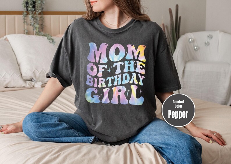 A woman wearing a black t-shirt with the text "MOM OF THE BIRTHDAY GIRL" printed on it, sitting on a bed.
