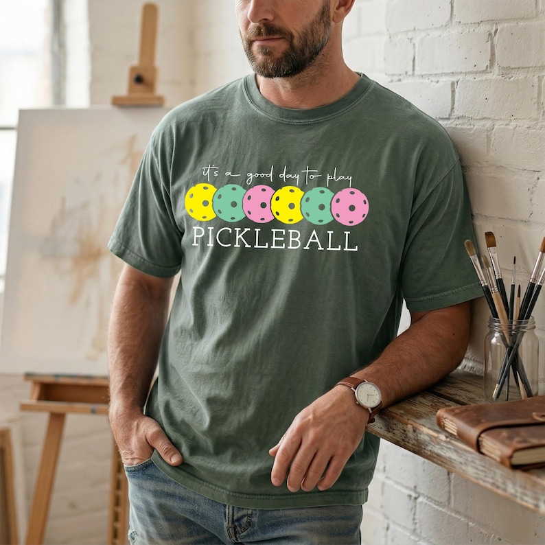 A man wearing a green t-shirt with a design of colorful pokckered balls and the text "It's a good day to play PICKLEBALL" is standing in front of a painting and a wooden easel.