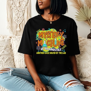 May include: Black t-shirt with a colorful graphic that reads "MYSTERY MACHINE of DIAGNOSTICS, ANOTHER CASE SOLVED BY THE LAB." The design features a beaker and other science-themed elements in orange, yellow, and blue. The person is wearing blue jeans.