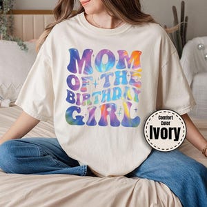 A woman wearing a white t-shirt with a colorful graphic design sitting on a bed.