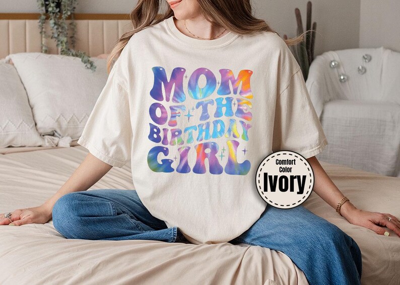 A woman wearing a white t-shirt with a colorful graphic design sitting on a bed.