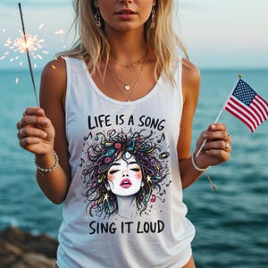 May include: A white sleeveless tank top featuring a graphic print of a stylised face with colourful, messy hair, musical notes, and vinyl records. The text "LIFE IS A SONG" appears above the graphic, and "SING IT LOUD" is printed below the illustration.