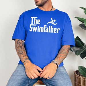 May include: Royal blue t-shirt with the text "The Swimfather" in white. The graphic includes a swimmer icon. The shirt is a casual wear item.