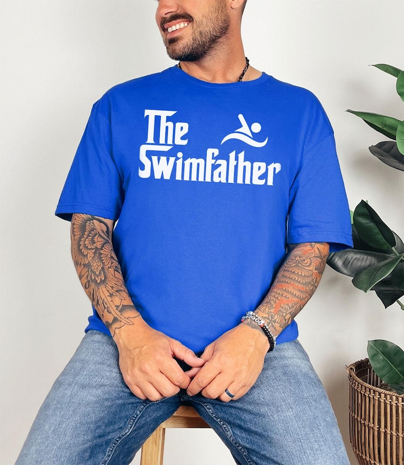 May include: Royal blue t-shirt with the text "The Swimfather" in white. The graphic includes a swimmer icon. The shirt is a casual wear item.