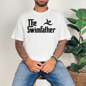 May include: White t-shirt with the text "The Swimfather" in black, featuring a swimmer icon. The shirt is a casual, short-sleeved design.