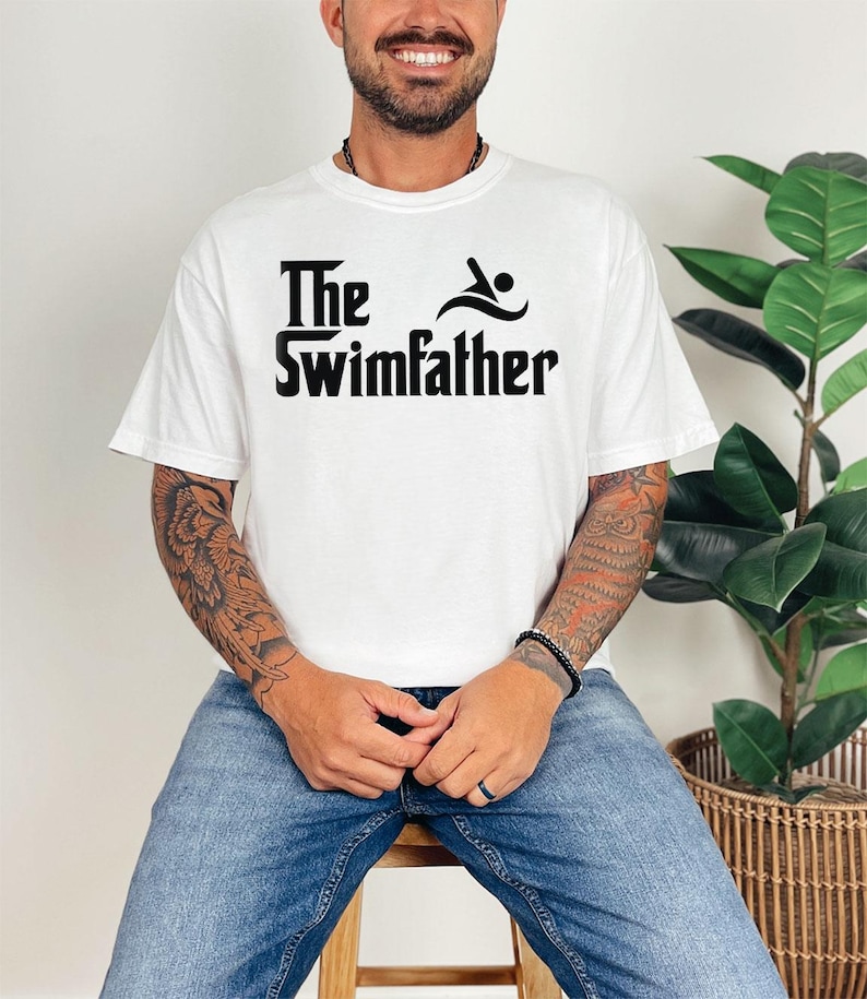 May include: White t-shirt with the text "The Swimfather" in black, featuring a swimmer icon. The shirt is a casual, short-sleeved design.