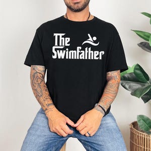 May include: Black t-shirt with the white text "The Swimfather" and a swimmer graphic. The shirt is made of a soft material and is suitable for casual wear. The design is centered on the chest.