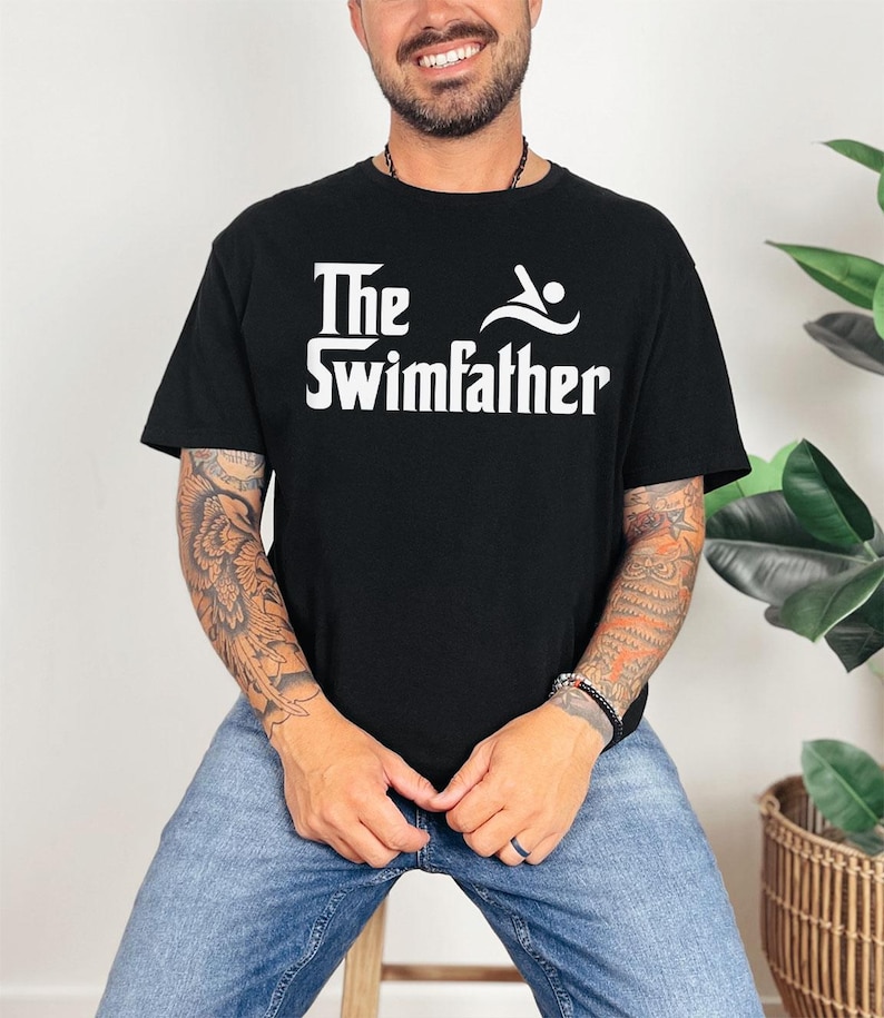 May include: Black t-shirt with the white text "The Swimfather" and a swimmer graphic. The shirt is made of a soft material and is suitable for casual wear. The design is centered on the chest.