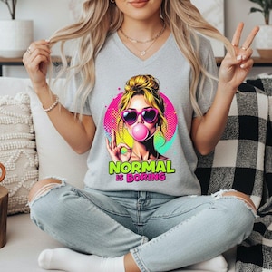May include: A heather gray V-neck t-shirt featuring a graphic of a person with blonde hair wearing heart-shaped sunglasses and blowing a pink bubble. The design includes a colorful circular background and the text "NORMAL IS BORING" in green and pink.