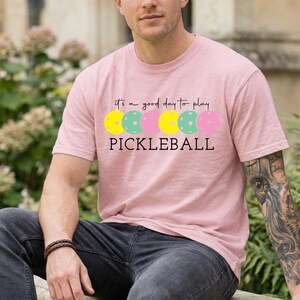 A man wearing a pink t-shirt with a graphic design of colorful pumas and the text "It's a good day to play PICKLEBALL" is sitting in a garden setting.