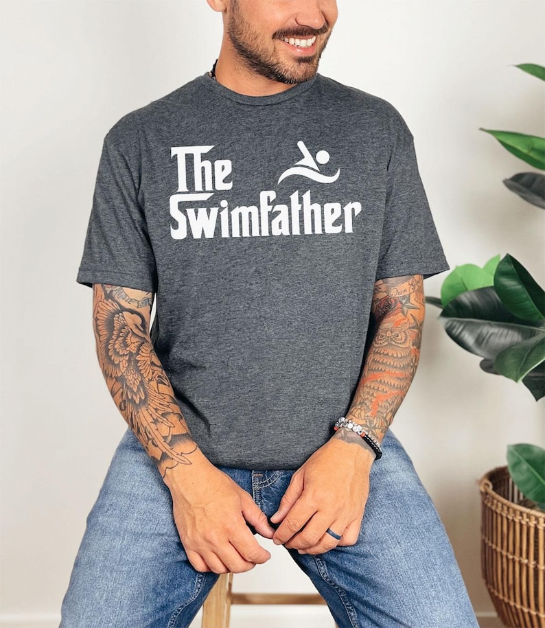 May include: Dark gray t-shirt with the text "The Swimfather" in white, featuring a swimmer graphic. The shirt is made of a soft material and is suitable for casual wear. The person in the image is wearing blue jeans.
