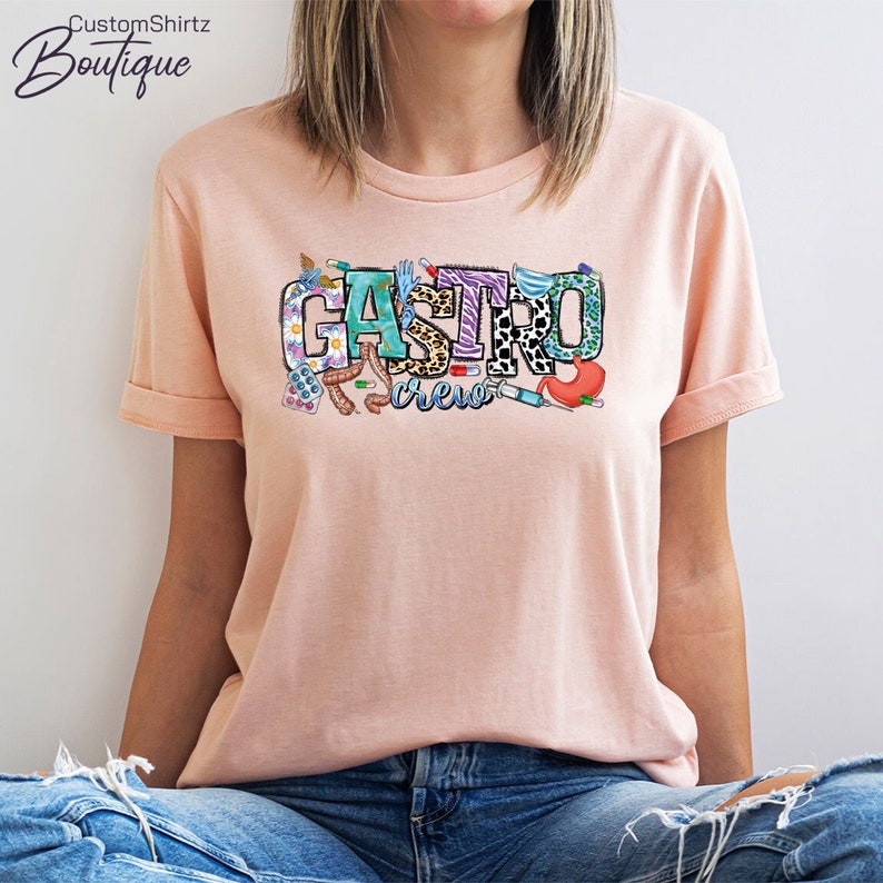 K&ouml;nnte beinhalten: Ein pinkfarbenes T-Shirt mit dem Text "Gastro Crew" in bunten Buchstaben mit verschiedenen medizinischen und gastrointestinalen Grafiken.
