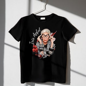 May include: Black t-shirt with a white graphic print of Iris Apfel wearing glasses and a patterned jacket. The graphic is surrounded by red and white paint splatters.