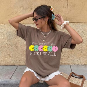 A woman sitting on a bench, wearing a brown t-shirt with a graphic of a pickleball and the text "It's a good day to play PICKLEBALL".