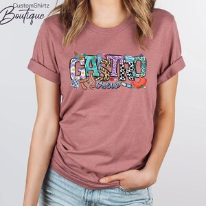 K&ouml;nnte beinhalten: Ein mauvefarbenes T-Shirt mit dem Text "Gastro Crew" in bunten Buchstaben. Die Buchstaben sind mit verschiedenen medizinischen Bildern verziert, darunter Pillen, ein Stethoskop und eine Spritze.
