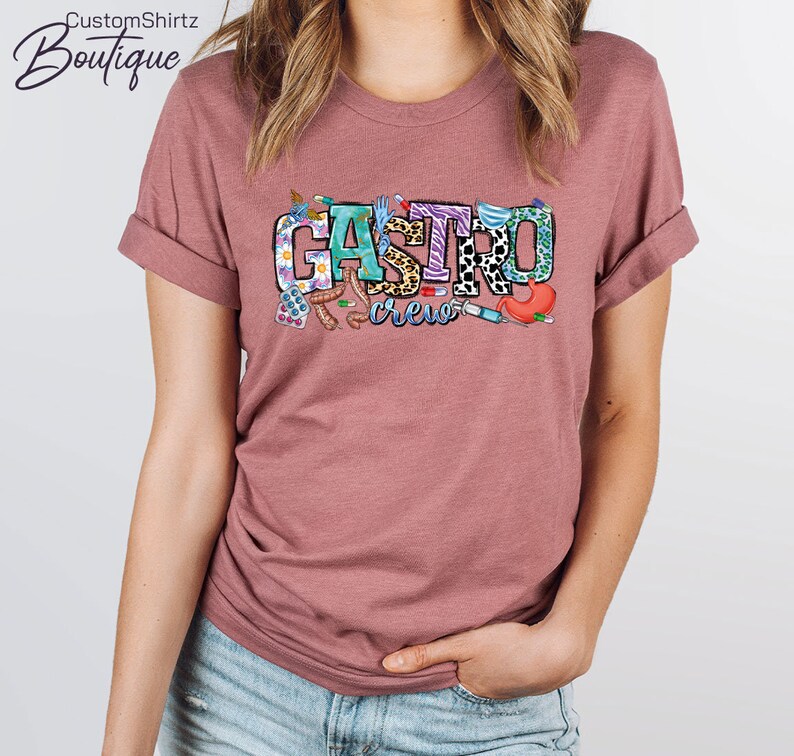K&ouml;nnte beinhalten: Ein mauvefarbenes T-Shirt mit dem Text "Gastro Crew" in bunten Buchstaben. Die Buchstaben sind mit verschiedenen medizinischen Bildern verziert, darunter Pillen, ein Stethoskop und eine Spritze.