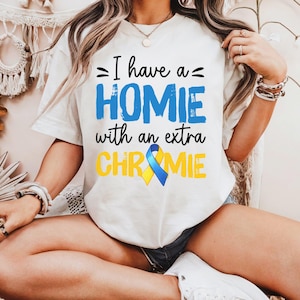 May include: White t-shirt featuring the text "I have a HOMIE with an extra CHROMIE" in blue and black. A blue and yellow ribbon graphic is below the text. The t-shirt is a casual style.