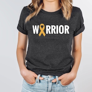 May include: A dark grey t-shirt with the word "WARRIOR" printed in white. A yellow ribbon is incorporated into the word "WARRIOR".