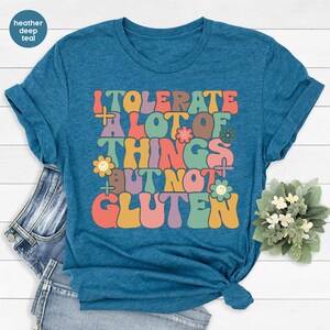 May include: A teal blue t-shirt with a retro-style graphic that says "I tolerate a lot of things but not gluten". The graphic is in rainbow colors with flowers and smiley faces.