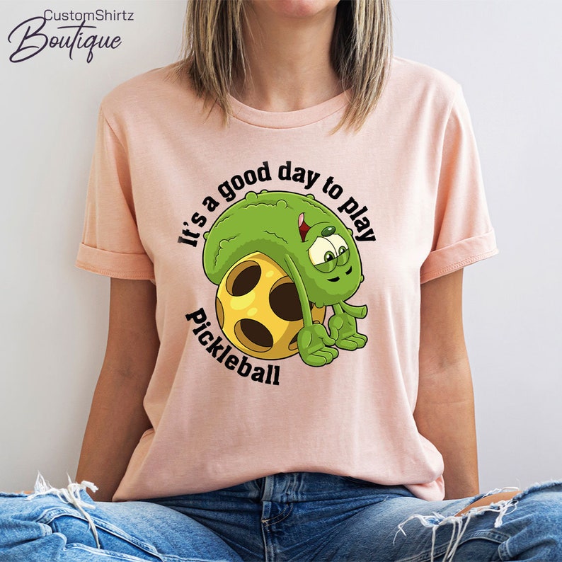 May include: A pink t-shirt with a green cartoon pickle character holding a yellow pickleball. The text on the shirt reads "It's a good day to play pickleball."