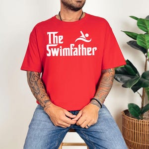 May include: A red t-shirt with the white text "The Swimfather" and a swimmer graphic. The shirt is short-sleeved and made of a soft material. The design is centered on the chest.