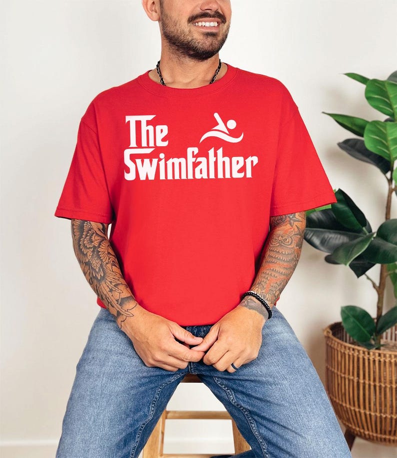 May include: A red t-shirt with the white text "The Swimfather" and a swimmer graphic. The shirt is short-sleeved and made of a soft material. The design is centered on the chest.