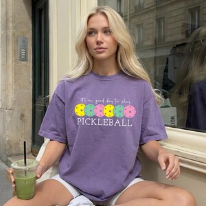 A young woman wearing a purple t-shirt with a playful design of colorful pokleball balls, sitting on a bench and holding a drink.