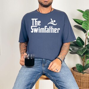 May include: Navy blue t-shirt with the text "The Swimfather" in white, featuring a swimmer graphic. The shirt is worn with blue jeans. A clear glass with a dark liquid is held in the hand.