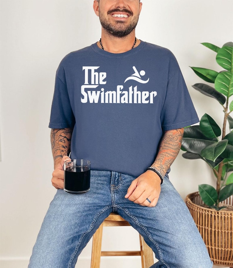 May include: Navy blue t-shirt with the text "The Swimfather" in white, featuring a swimmer graphic. The shirt is worn with blue jeans. A clear glass with a dark liquid is held in the hand.