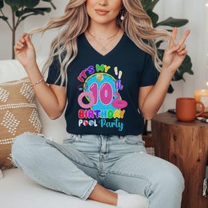 May include: Navy blue v-neck t-shirt with a colorful graphic that reads "It's My 10th Birthday Pool Party." The graphic includes a pink inflatable flamingo, a pink inflatable donut, and other pool-themed elements. The shirt is a birthday party theme.