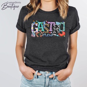 K&ouml;nnte beinhalten: Ein dunkelgraues T-Shirt mit dem Text "Gastro crew" in bunten Buchstaben. Die Buchstaben sind mit verschiedenen medizinischen Bildern verziert, darunter Pillen, ein Stethoskop, eine Spritze und ein Magen.