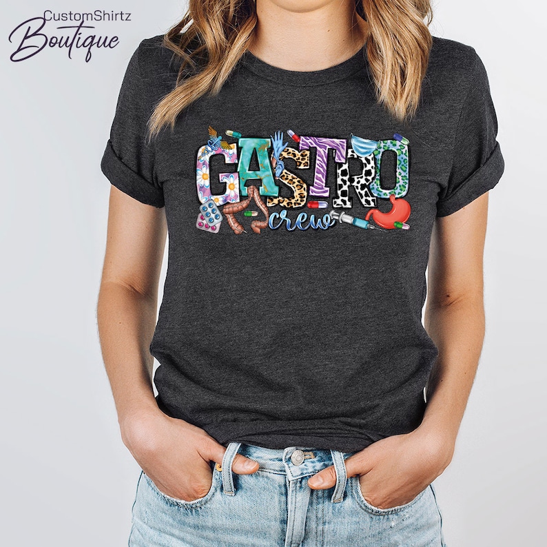 K&ouml;nnte beinhalten: Ein dunkelgraues T-Shirt mit dem Text "Gastro crew" in bunten Buchstaben. Die Buchstaben sind mit verschiedenen medizinischen Bildern verziert, darunter Pillen, ein Stethoskop, eine Spritze und ein Magen.