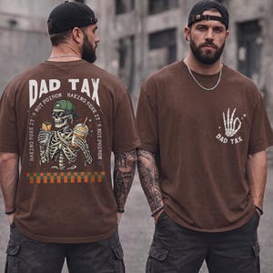 May include: A brown t-shirt featuring a graphic on the back of a skeleton wearing a green cap and sunglasses, holding a food item and a drink, with the text DAD TAX. The front features a smaller graphic of a skeletal hand making an okay sign.