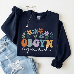 May include: Navy blue sweatshirt with the text "OBGYN squad" in colorful letters and a floral design. The sweatshirt is paired with blue jeans and a necklace.