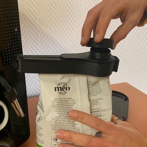 May include: A black coffee bag sealer is being used to close a white bag of coffee. The bag has the word "méo" printed on it. The sealer is attached to a coffee machine.