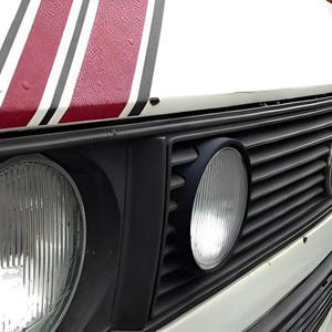 Peut inclure: Un fourgon blanc avec une bande noire et rouge sur le capot. Le fourgon a une calandre noire avec un phare rond sur le côté droit. La calandre porte le logo Volkswagen.