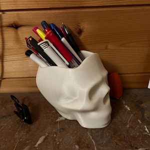 Peut inclure: Organiseur de bureau en forme de crâne imprimé en 3D blanc avec une variété de stylos et de crayons à l'intérieur.