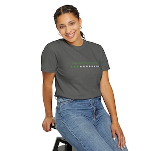 Puede incluir: Una camiseta gris con el texto "You Are Now In Line" en verde y blanco. La camiseta está hecha de un material suave y la lleva una persona.
