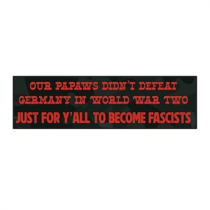 May include: A rectangular sign with a black and green camouflage background. Bold red text reads: "OUR PAPAWS DIDN'T DEFEAT GERMANY IN WORLD WAR TWO JUST FOR Y'ALL TO BECOME FASCISTS."