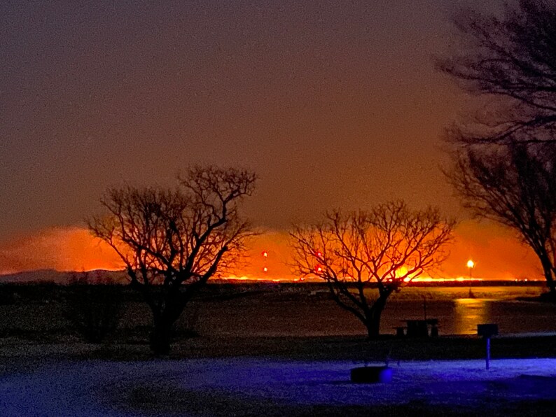 K&ouml;nnte beinhalten: Eine Nachtaufnahme eines Sees mit einer leuchtend orangefarbenen Skyline einer Stadt im Hintergrund. Im Vordergrund sind Silhouetten von B&auml;umen zu sehen, in der Ferne ein Picknicktisch und ein Grill.