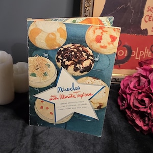 May include: A vintage cookbook titled Miracles with Minute Tapioca featuring a blue cover with a white star graphic and illustrations of various tapioca desserts in glass bowls. The book is surrounded by dark red roses and white candles.