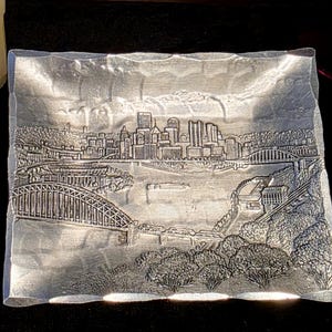 May include: A rectangular silver-coloured metal tray featuring a raised embossed design of a city skyline with multiple bridges spanning a river. The tray has a textured, hammered-style border and a detailed depiction of urban buildings and foliage.