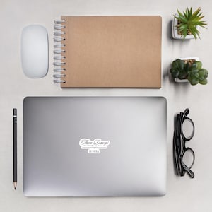 May include: A laptop sticker with the text "Them Dawgs is Hell" in a cursive font. The sticker is on a silver laptop, with a mouse, notebook, pencil, glasses, and small plants nearby.